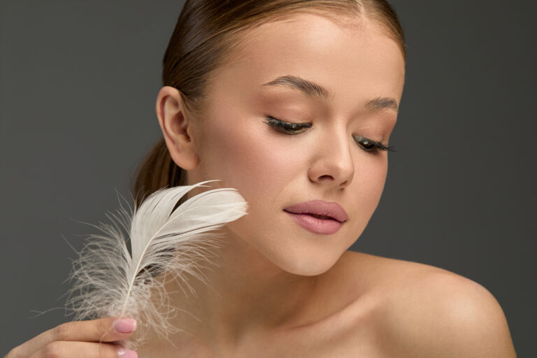 A woman gently touches her cheek with a soft feather while applying lotion. She showcases her smooth skin and natural beauty in a serene environment.