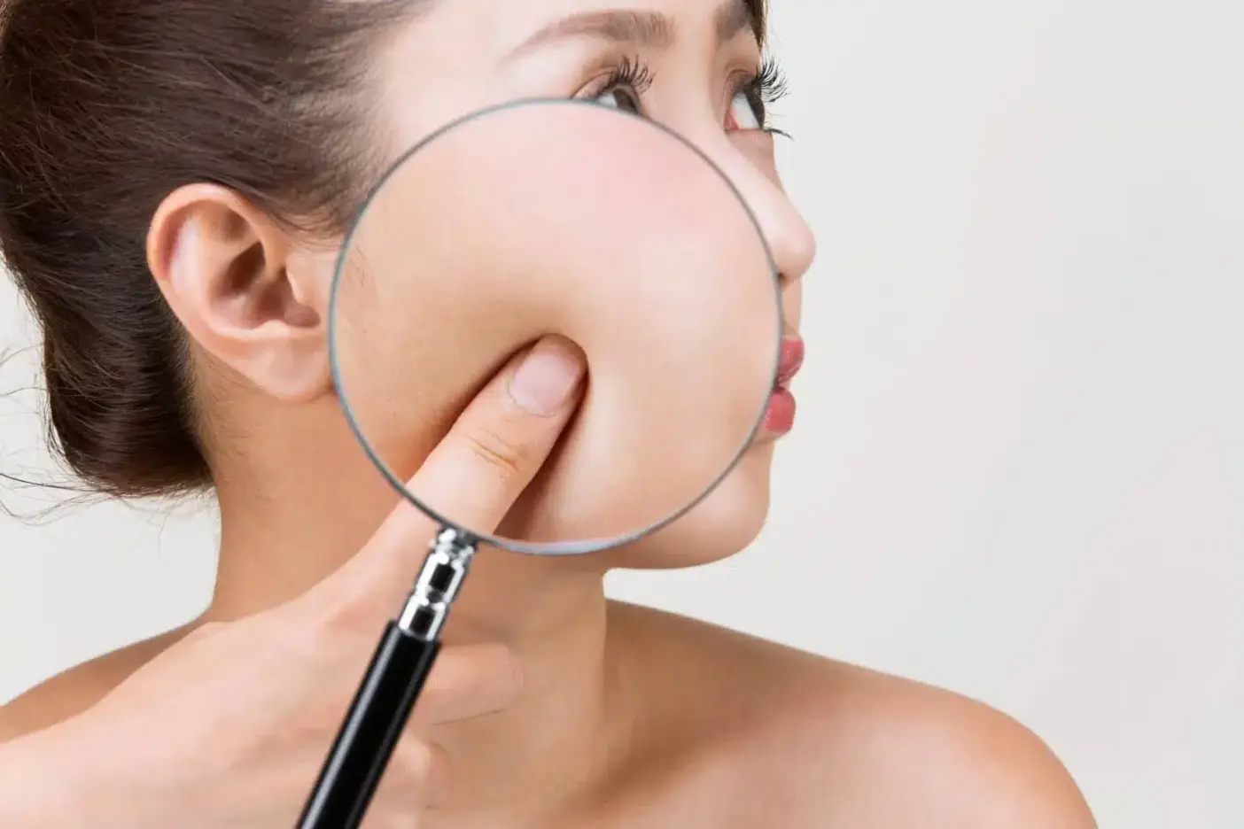 A woman holds a magnifying glass to her face and examines her skin.