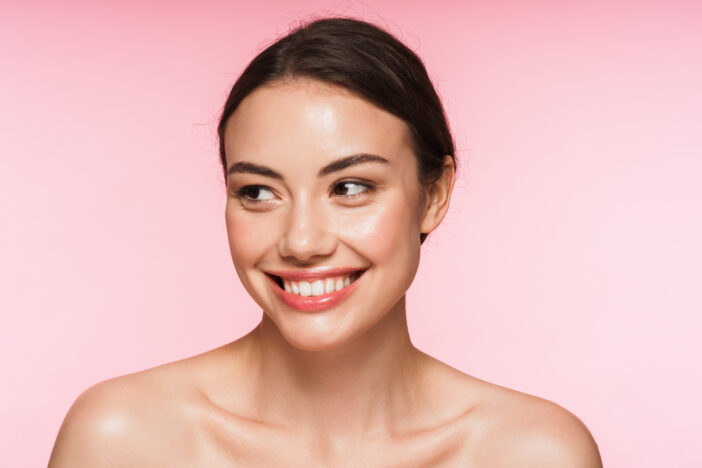 Beauty portrait of a beautiful smiling young brunette woman with glowing skin standing isolated over pink background